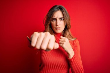 Genç ve güzel bir esmer kadın, kırmızı arka planda balıkçı yaka kazak giyiyor. Yumruğunu yumrukluyor, saldırıyor, saldırıyor, tehdit ediyor ve şiddet uyguluyor.