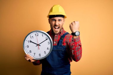 Güvenlik kaskı takan genç adam sarı arka planda büyük bir saat tutuyor. Sinirli ve sinirli bir şekilde bağırıyor, çıldırıyor ve el kaldırarak bağırıyor. Öfke kavramı.