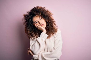 Kıvırcık saçlı ve piercingli güzel bir kadın pembe arka planda sıradan bir süveter giyiyor. Kollarını kavuşturup depresyondan sıkılmış ve yorgun görünüyor..
