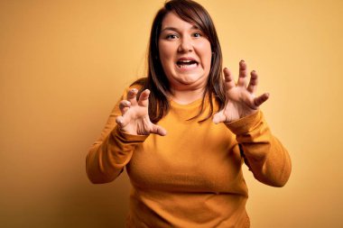 Güzel esmer ve cüsseli bir kadın izole edilmiş sarı arka planda rahat bir süveter giyiyor komik bir şekilde kedi gibi pençe hareketi yapıyor, agresif ve seksi bir ifade.