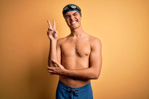 若いハンサムな男は 水着を着て 指で勝利サインを行うカメラでの幸せな顔のウィンクで笑顔孤立した黄色の背景の上にキャップを泳ぐ 第二番 — ストック写真