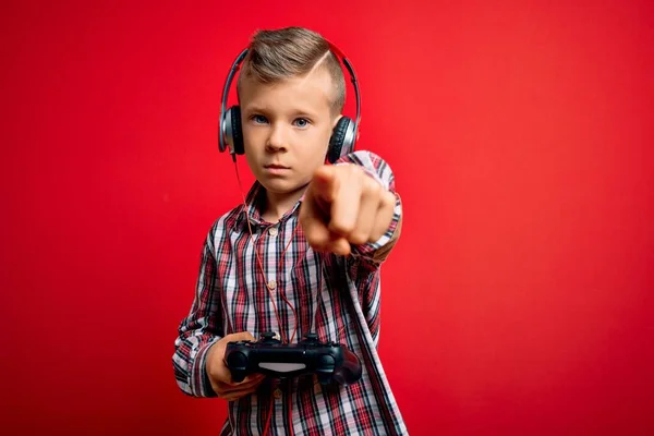 若い小さな白人のゲーマーの子供は カメラとあなたに指で指している赤い背景にゲームパッドジョイスティックを使用してビデオゲームをプレイします 手のサイン 正とフロントからの自信を持ってジェスチャー — ストック写真