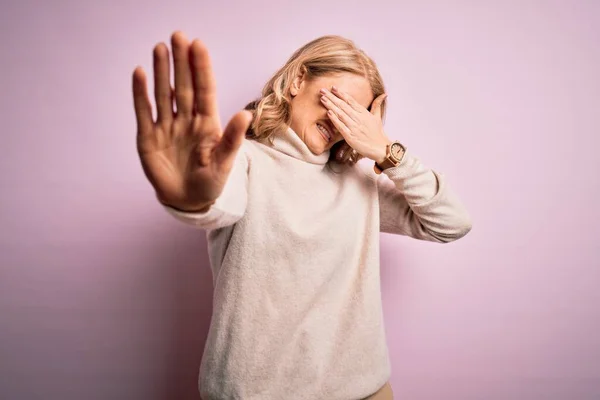 Middle age beautiful blonde woman wearing casual turtleneck sweater over pink background covering eyes with hands and doing stop gesture with sad and fear expression. Embarrassed and negative concept.