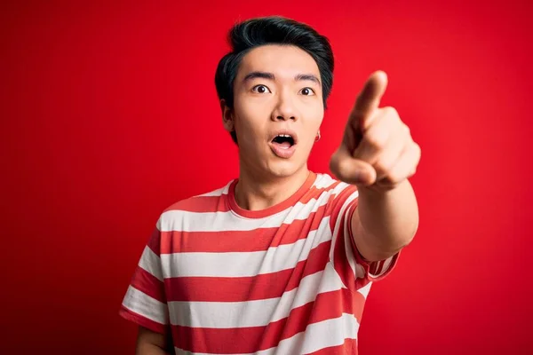 若いハンサムな中国人男性のカジュアルな縞模様のTシャツの赤い背景の上に立って指を前に 口を開けて驚いて表現 フロントに何かを指して — ストック写真