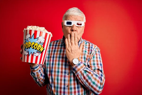 Senior hoary man watching film using 3d glasses eating popcorn over red background cover mouth with hand shocked with shame for mistake, expression of fear, scared in silence, secret concept