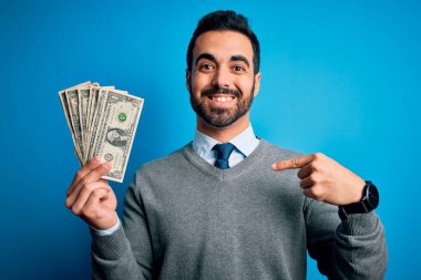 Sakallı, mavi arka planda banknotlar tutan yakışıklı genç bir adam.