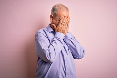 Orta yaşlı yakışıklı, beyaz bir adam pembe arka planda dikilmiş ağlarken suratını ellerle kapatan sıradan bir gömlek giyiyor. Depresyon kavramı.