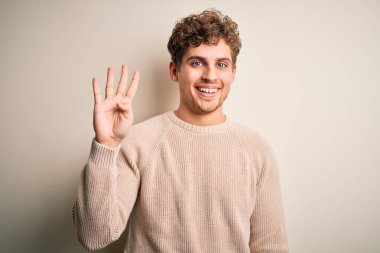 Sarışın, kıvırcık saçlı, beyaz arka plan üzerine günlük süveter giyen, dört numarayı işaret eden, kendinden emin ve mutlu gülümseyen genç bir adam..