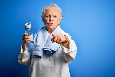 Üst düzey, güzel, spor bir kadın elinde bir şişe suyla izole edilmiş mavi arka planda dikilip kamerayı işaret ediyor ve sen, el işareti, ön taraftan pozitif ve kendinden emin bir hareket.