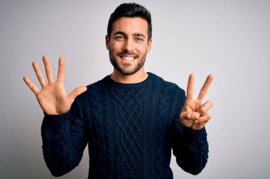 Sakallı, sıradan bir süveter giyen, beyaz arka plan üzerinde duran ve yedi numaralı parmaklarıyla işaret eden genç ve yakışıklı bir adam kendinden emin ve mutlu bir şekilde gülümsüyor..