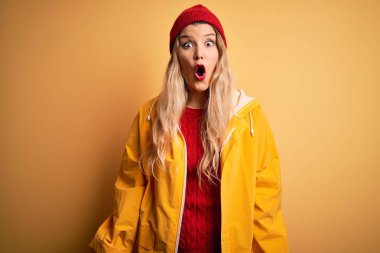 Sarışın, yağmurluk ve yünlü şapka giyen genç ve güzel bir kadın izole edilmiş sarı arka planda korku, korku ve heyecan dolu bir yüz ifadesiyle dehşete düşmüş ve şok olmuştu..