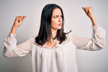 Mavi gözlü genç esmer kadın izole edilmiş beyaz arka plan üzerinde sıradan tişörtler giyiyor ve gururla gülümseyen kol kaslarını gösteriyor. Fitness konsepti.