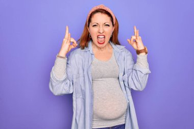 Güzel, kızıl saçlı hamile bir kadın, izole edilmiş mor arka planda bebek bekliyor. Bağırıyor, çılgın bir ifadeyle elleri havada rock sembolü yapıyor. Müzik yıldızı. Ağır konsept.
