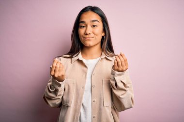 Genç, güzel Asyalı bir kadın pembe arka planda dikilmiş para jestleri yapıyor, maaş istiyor, milyonerlik işi yapıyor.