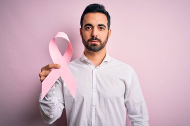 Sakallı, pembe kanser kurdelesi takan genç, yakışıklı bir adam izole bir arka planda, akıllı yüzünde kendine güvenen bir ifadeyle ciddi düşünüyor.