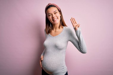 Genç ve güzel bir kız hamile, pembe arka planda bebek bekliyor. Üç numaralı parmaklarıyla işaret ediyor. Kendinden emin ve mutlu bir şekilde gülümsüyor..