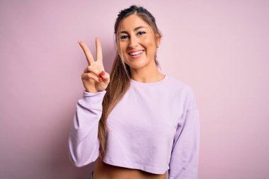 Pembe izole edilmiş arka plan üzerinde eşofman üstü giyen genç ve güzel bir bayan iki numaralı parmaklarıyla gülümseyerek kendinden emin ve mutlu bir şekilde onu işaret ediyor..