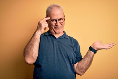 Sarı arka plan üzerine gündelik polo ve gözlük takan orta yaşlı, beyaz bir adam. Avuç içinin açık olması, fotokopi alanını göstermesi ve alnını işaret etmesi kafasını karıştırmış. Bunu bir düşün..