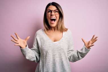 Genç, güzel, esmer bir kadın pembe arka plan üzerinde gözlük ve süveter takıyor deli deli deli bağırıyor ve agresif bir ifade ve kollar havada bağırıyor. Hayal kırıklığı konsepti.
