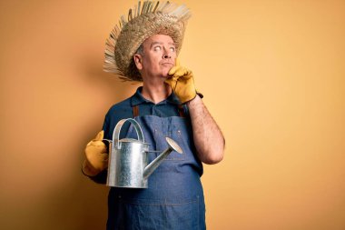 Orta yaşlı çiftçi, önlük ve şapka takıyor. Sarı arka plan üzerinde su kutusu var. Ciddi bir yüz.