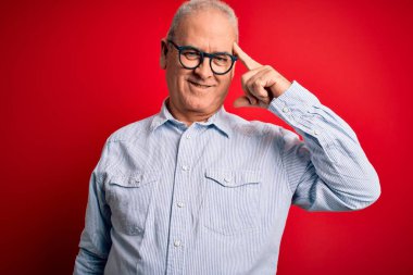 Orta yaşlı, yakışıklı, beyaz gömlekli ve kırmızı arka planda gözlüklü bir adam. Tek parmağıyla kafasını gösteren, harika bir fikir ya da düşünce, iyi bir hafıza.