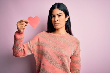 Genç, güzel, İspanyol bir kadın pembe arka planda romantik bir kalp kâğıdı tutuyor. Yüzünde kendine güvenen bir ifade var.