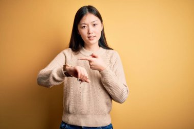 Sarı izole edilmiş arka plan üzerine sıradan süveter giyen genç, güzel Asyalı bir kadın. Aceleyle saati, sabırsızlığı, gecikmeye kızmış ve sinirli bir şekilde izliyordu.