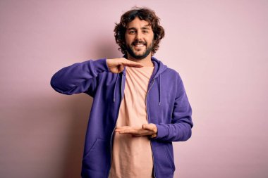 Sakallı, pembe arka planda sıradan bir eşofman üstü giyen, büyük ve büyük beden işareti olan genç, yakışıklı bir adam. Ölçü sembolü. Kameraya bakıp gülümsüyor. Ölçüm konsepti.