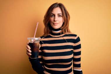 Genç, güzel, esmer bir kadın kola ile birlikte bardak içiyor. Saman kullanıyor. Kendine güvenen bir ifade takınıyor.