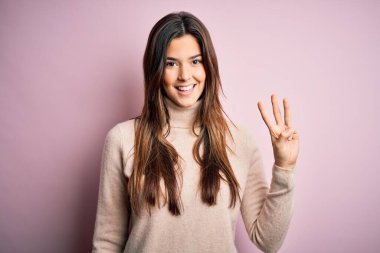 Balıkçı yaka kazak giyen genç ve güzel bir kız soyutlanmış pembe arka plan üzerinde duruyor ve üç numarayı işaret ederken kendinden emin ve mutlu bir şekilde gülümsüyor..