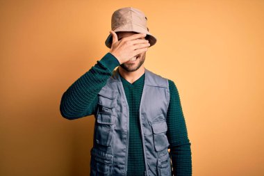 Tatilde sakallı yakışıklı turist sarı arka planda kaşif şapkası takıyor, gülümsüyor ve yüzünde şaşkınlıktan gözleri kapayarak gülüyor. Kör kavram.