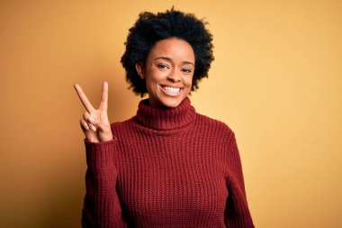 Kıvırcık saçlı, balıkçı yaka kazak giyen, gülümseyen, parmaklarını zafer işareti yaparken gösteren kameraya bakan güzel Afro-Amerikan kadın. İki numara..