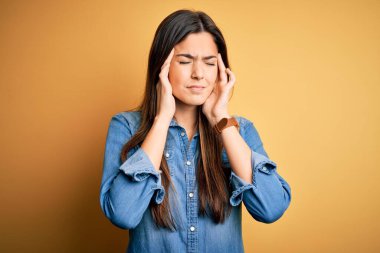 İzole edilmiş sarı arka plan üzerinde duran sıradan bir kot gömlek giyen genç ve güzel bir kız. Başı ağrıyor çünkü stres altında. Migren ağrısı..