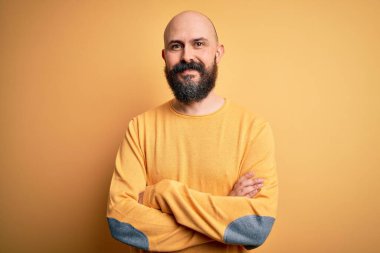 Sarışın, kel ve sakallı bir adam sarı arka planda dikilip kollarını kavuşturup kameraya bakarken gülümsüyor. Pozitif kişi.