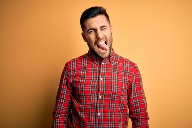 Genç yakışıklı adam rahat gömlek giyen izole sarı arka plan üzerinde duran komik ifade ile mutlu dil yapışmasını. Duygu kavramı.
