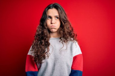 Kıvırcık saçlı genç bir kadın izole edilmiş kırmızı arka planda komik yüzlü, şişirilmiş yanakları olan günlük bir kazak giyiyor. Havayla şişmiş ağız, çılgın bir ifade..