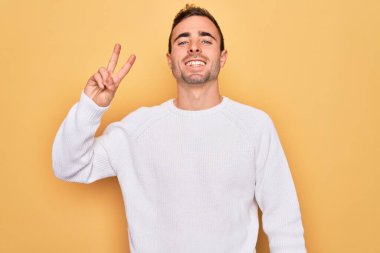 Mavi gözlü, sıradan bir süveter giyen, sarı arka planda durup gülümseyen ve parmaklarını zafer işareti yapan genç yakışıklı bir adam. İki numara..