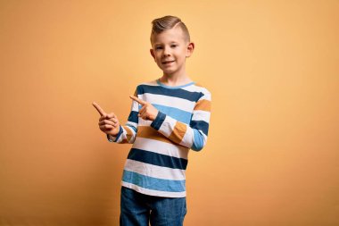 Küçük, beyaz, mavi gözlü, sarı arka planda renkli çizgili gömlek giyen, gülümseyen ve kameraya bakıp iki eliyle ve parmaklarıyla yüzünü gösteren bir çocuk..