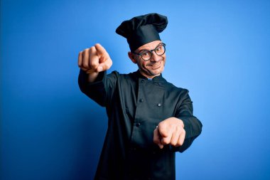 Genç, yakışıklı aşçı üniformalı ve şapkalı, izole edilmiş mavi arka planda seni ve kamerayı parmaklarıyla gösteren neşeli ve pozitif gülümseyen bir aşçı.