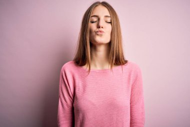 Genç, güzel, kızıl saçlı bir kadın pembe arka planda rahat bir süveter giyiyor kameraya bakıyor ve güzel ve seksi bir şekilde havada öpüşüyor. Aşk ifadesi.