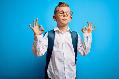 Mavi arka planda akıllı gözlüklü ve okul çantalı küçük beyaz çocuk rahatla ve gözleri kapalı meditasyon hareketi yaparken parmaklarınla gülümse. Yoga konsepti.