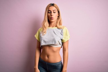 Genç ve güzel sarışın spor kadın spor yapıyor. Pembe arka planda spor kıyafetleri giyiyor. Dudaklı balık suratı yapıyor, çılgınca ve komik jestler yapıyor. Komik ifade.