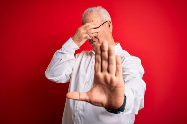 Orta yaşlı yakışıklı, beyaz gömlekli, gözlüklü, kırmızı arka planda gözlüklü, gözleri ellerle kaplayan, üzgün ve korku ifadeli hareketler yapan bir adam. Utanç verici ve olumsuz bir kavram..