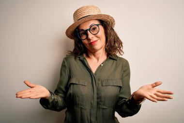 Orta yaşlı esmer kadın, gözlüklü ve şapkalı, soyutlanmış beyaz arka planda duruyor. Kolları ve elleri havada, kafası karışmış bir ifade. Şüphe kavramı.