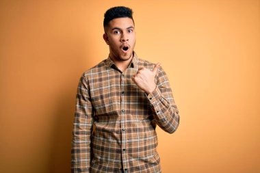 İzole edilmiş sarı arka planda dikilen sıradan tişörtlü genç ve yakışıklı bir adam. El parmağı yana dönük, ağzı açık, şaşkın bir ifade..