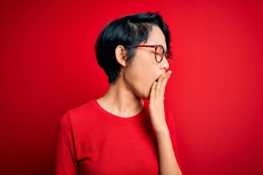 Genç, güzel Asyalı bir kız, gündelik tişört ve gözlük takıp izole edilmiş kırmızı arka planda esnemekten sıkılmış, ağzıyla ağzını kapatmaktan sıkılmış. Huzursuz ve uykulu..