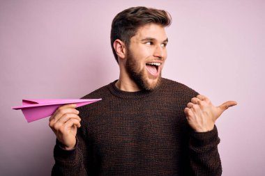 Sarışın, sakallı, mavi gözlü, pembe kâğıt uçakla arka planda gezen ve gülümseyen bir yüzü olan genç bir adam.