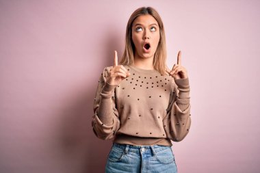 Pembe izole edilmiş arka planda güzel sarışın kadın şaşırmış ve şaşırmış bir şekilde yukarı bakıp parmaklarıyla işaret ediyor ve kollarını kaldırıyordu..