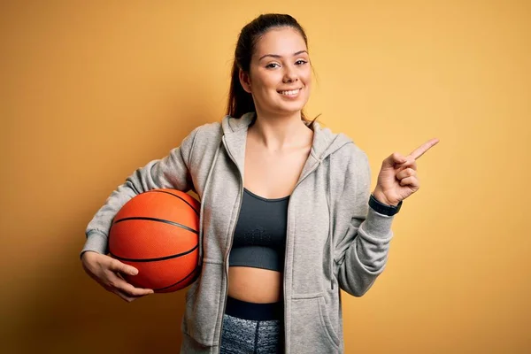 年轻美丽的黑发女运动员拿着黄色背景的篮球运动者非常高兴地将手和手指指向旁边 — 图库照片