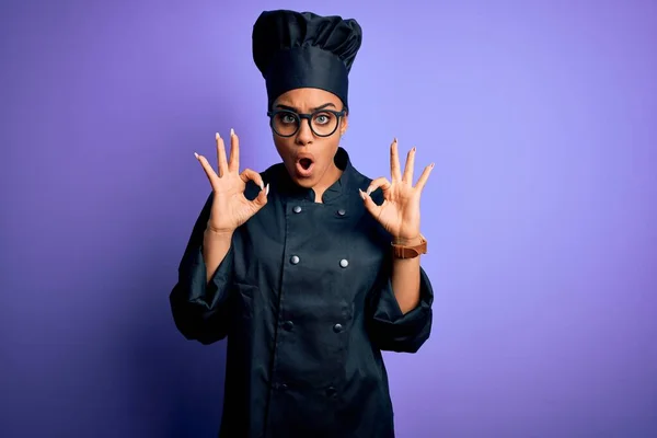 年轻的非洲女厨师身穿炊具制服 头戴紫色背景的帽子 看上去很惊讶 用手指做了个不错的认可标志 疯狂的表情 — 图库照片